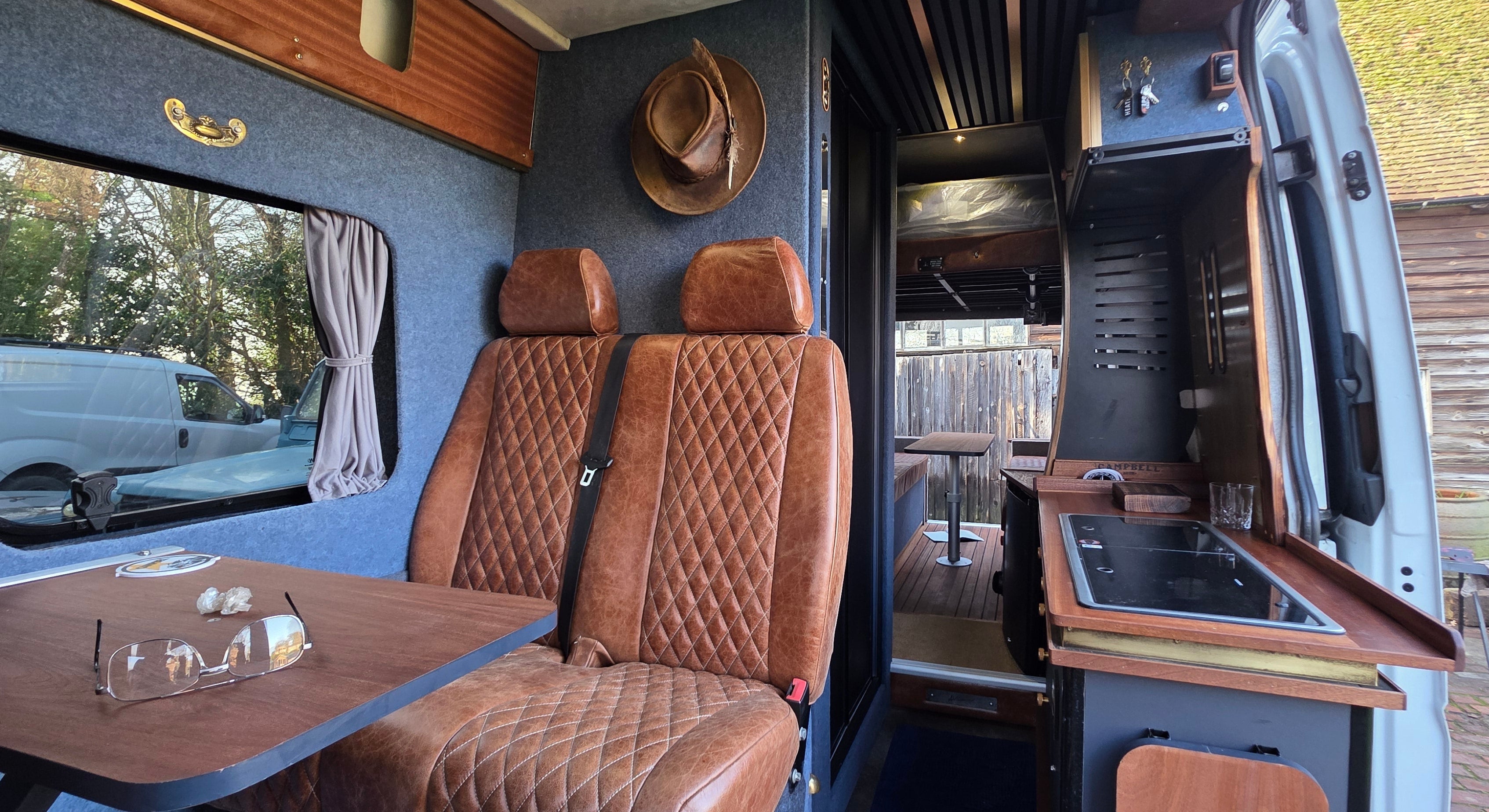 Interior of a van with brown seats, table, and kitchen area.
